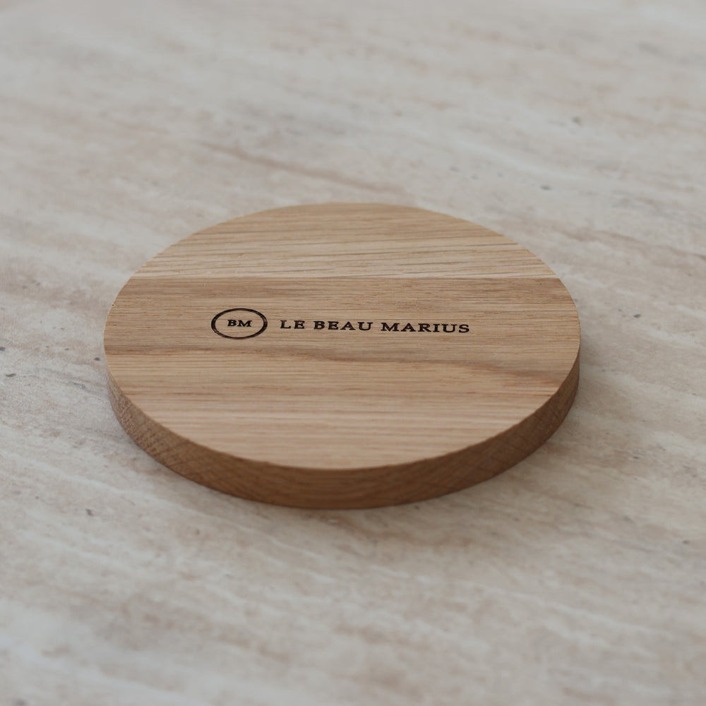 Dessous de mug en chêne massif Ø 10 cm gravé Le Beau Marius, posé sur du travertin